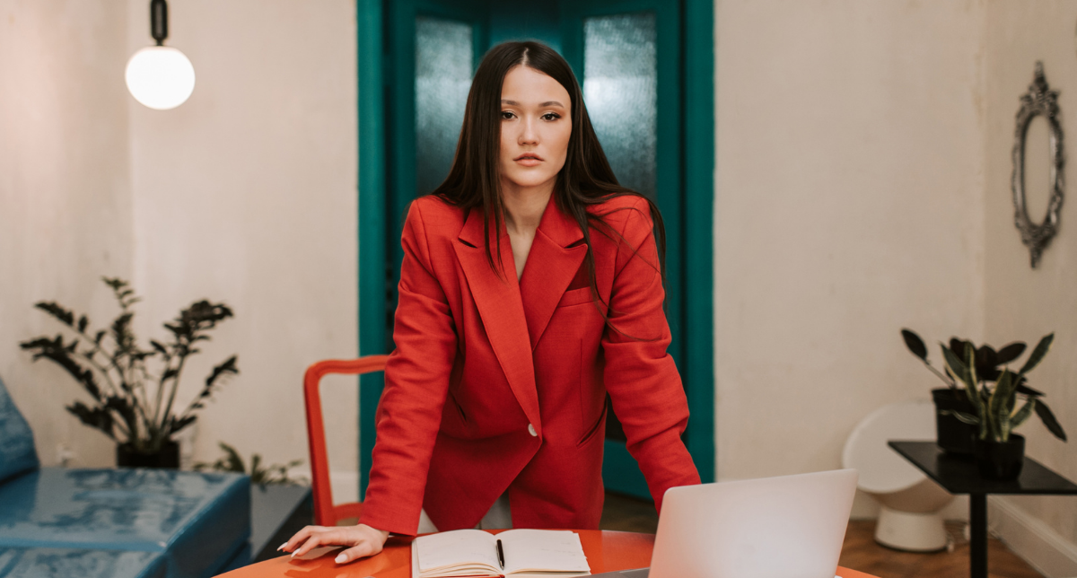 Kinavic Leadership Acceleration - Want Bold Leadership in the Era of AI? A young woman in a bold red suit stands over a work desk with her computer with a serious look on her face.